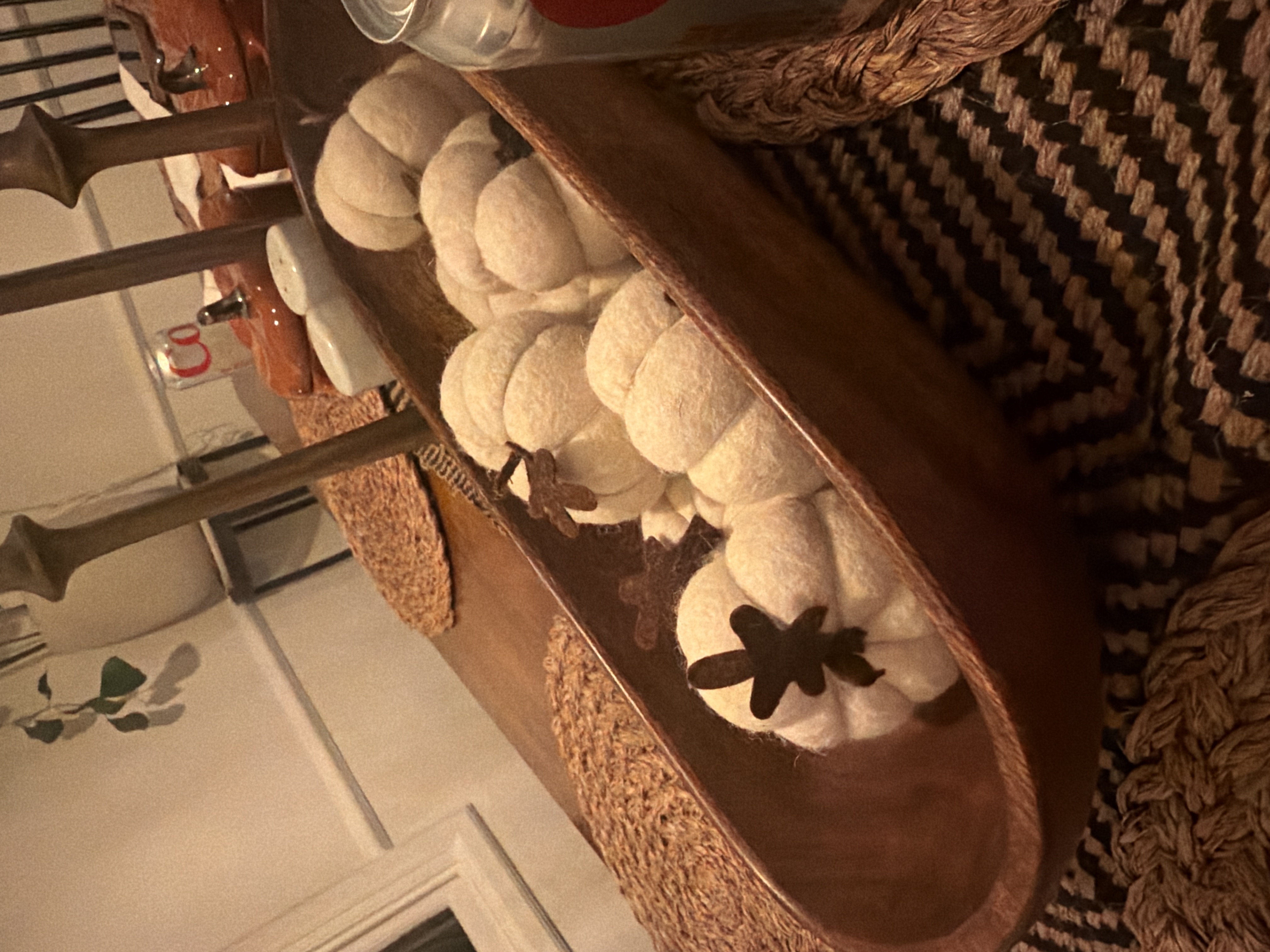 Soft cream fabric pumpkins with dark leaf accents nestled in a long wooden tray on a warmly lit dining table, the featured autumn centerpiece.