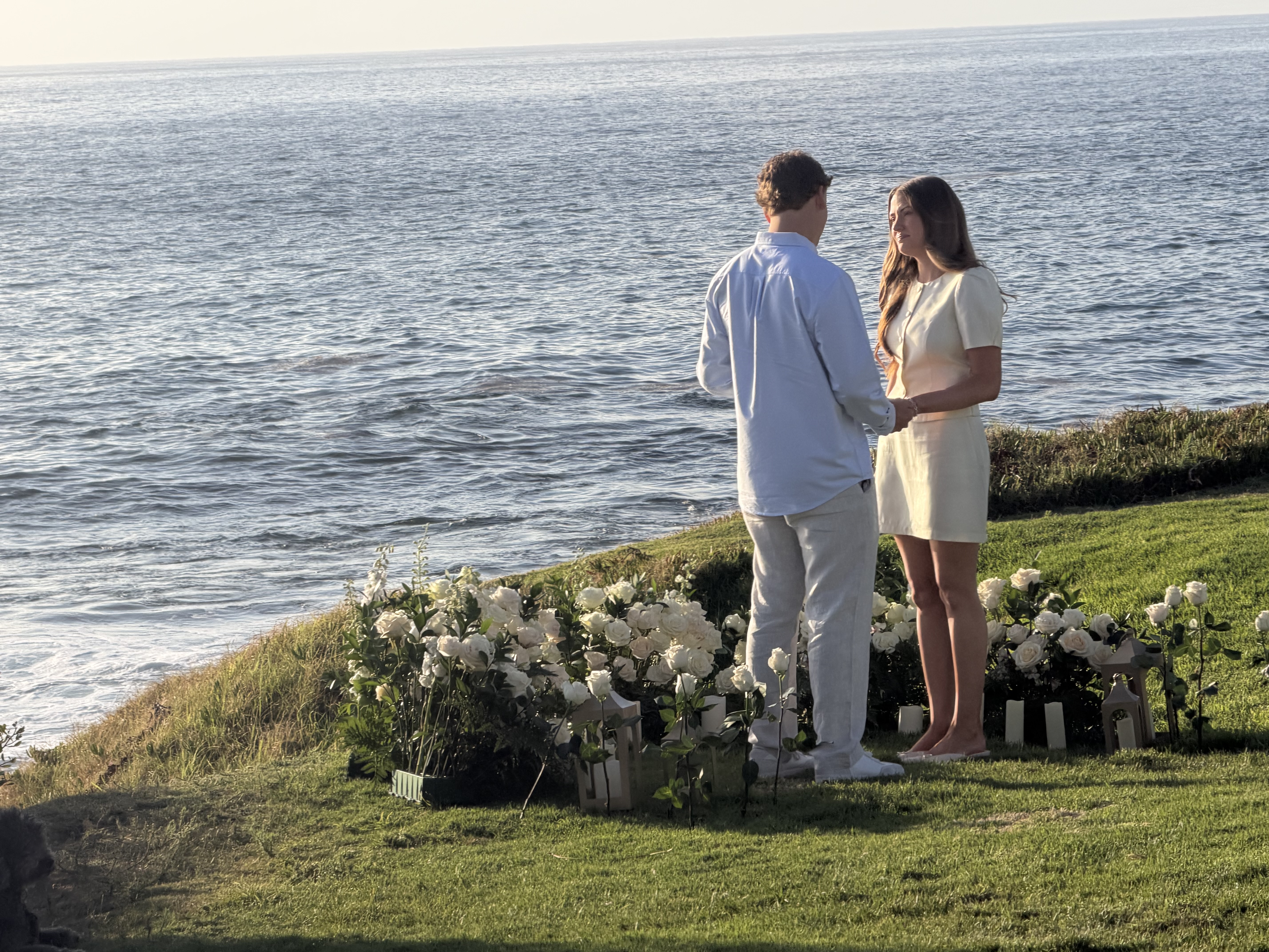Markus and Sam holding hands surrounded by white roses on a grassy La Jolla cliff overlooking the ocean at sunset